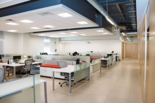 A modern office space featuring a row of cubicles with desks and office chairs. The area is well-lit with overhead lights and large windows. The flooring is tiled, and there are a few people, including an employment retaliation attorney, working diligently at their desks.